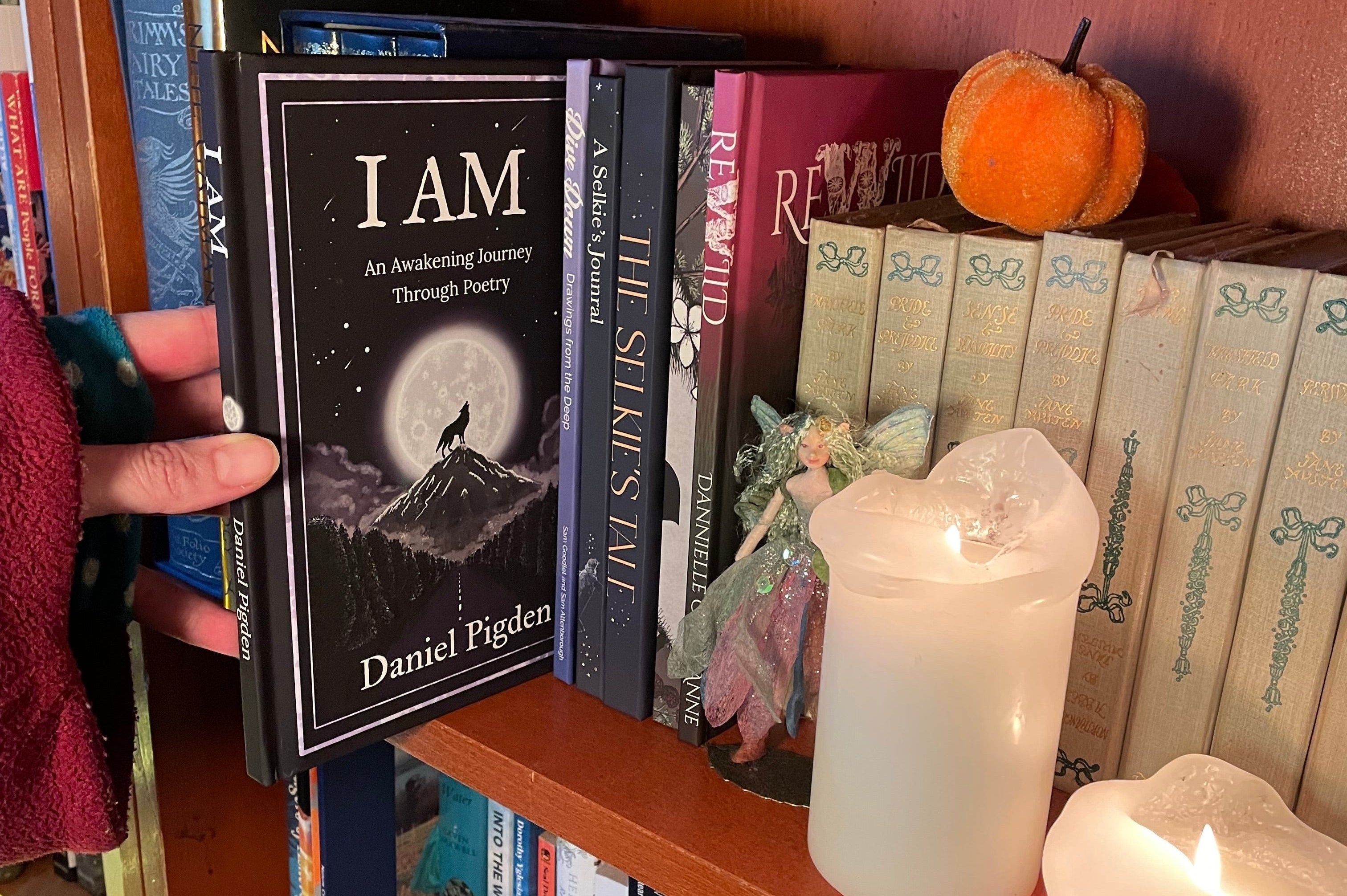 A hand holds a copy of the poetry book 'I AM: An Awakening Journey Through Poetry' by Daniel Pigden, standing next to other books from Wolf and Wing on a wooden shelf with a glowing white candle in the foreground.