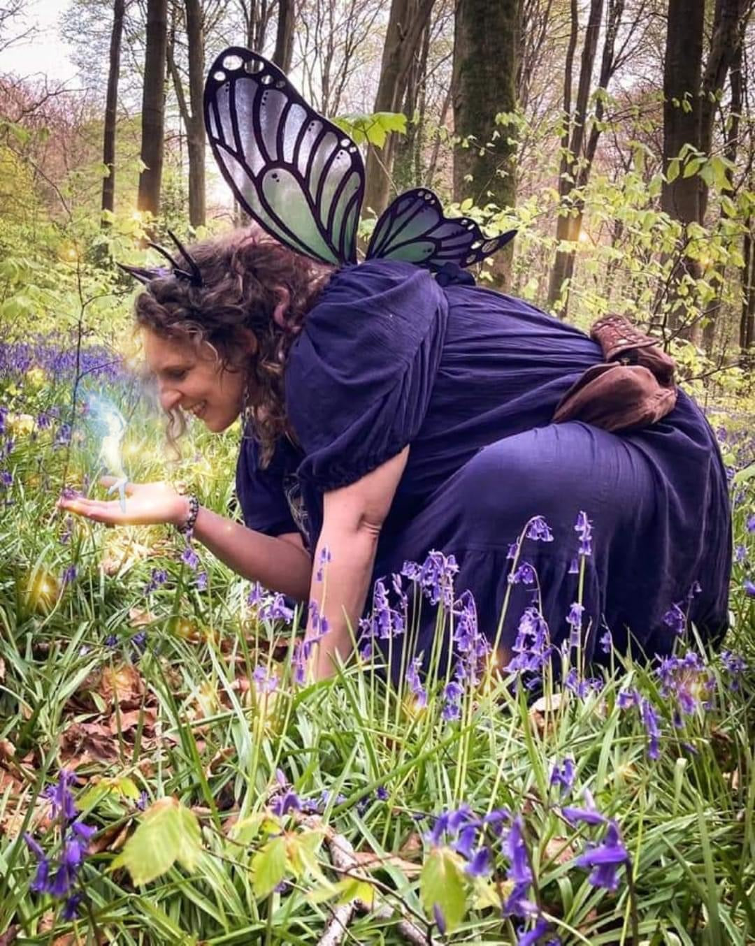 Sam wearing fairy wings crouches in a bluebell wood